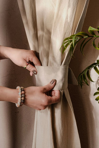 Hands fastening the tieback band of a beige stonewashed cotton curtain. Handwoven in Denizli, OEKO-TEX. Vivamaison.
